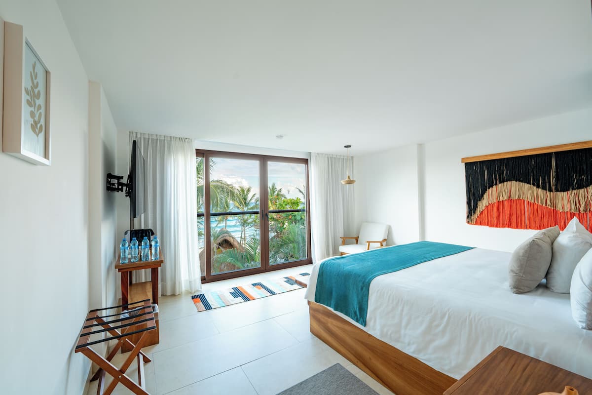 A modern hotel room with a large bed, a TV, a small table with water bottles, and a view of palm trees through the window.