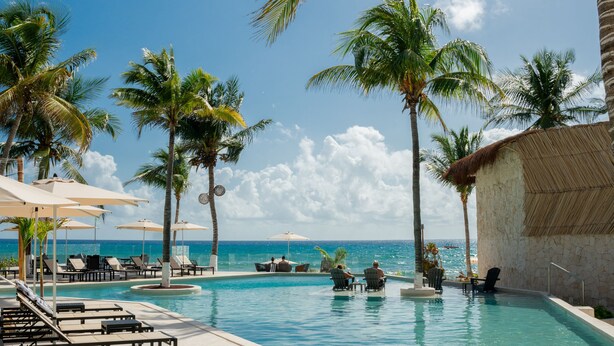 A clear blue pool with lounge chairs and umbrellas, surrounded by palm trees and a view of the ocean.