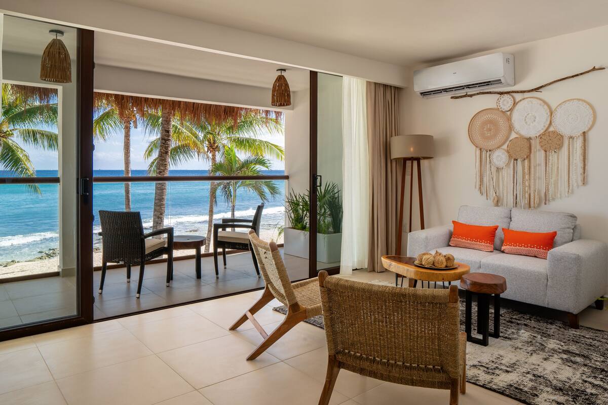 A modern living room with a balcony overlooking the ocean, featuring wicker furniture, a grey sofa with orange cushions, and a wall decorated with dreamcatchers.