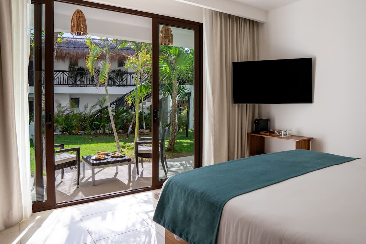 A bedroom with a bed, a TV, and a view of a balcony with outdoor furniture and plants.