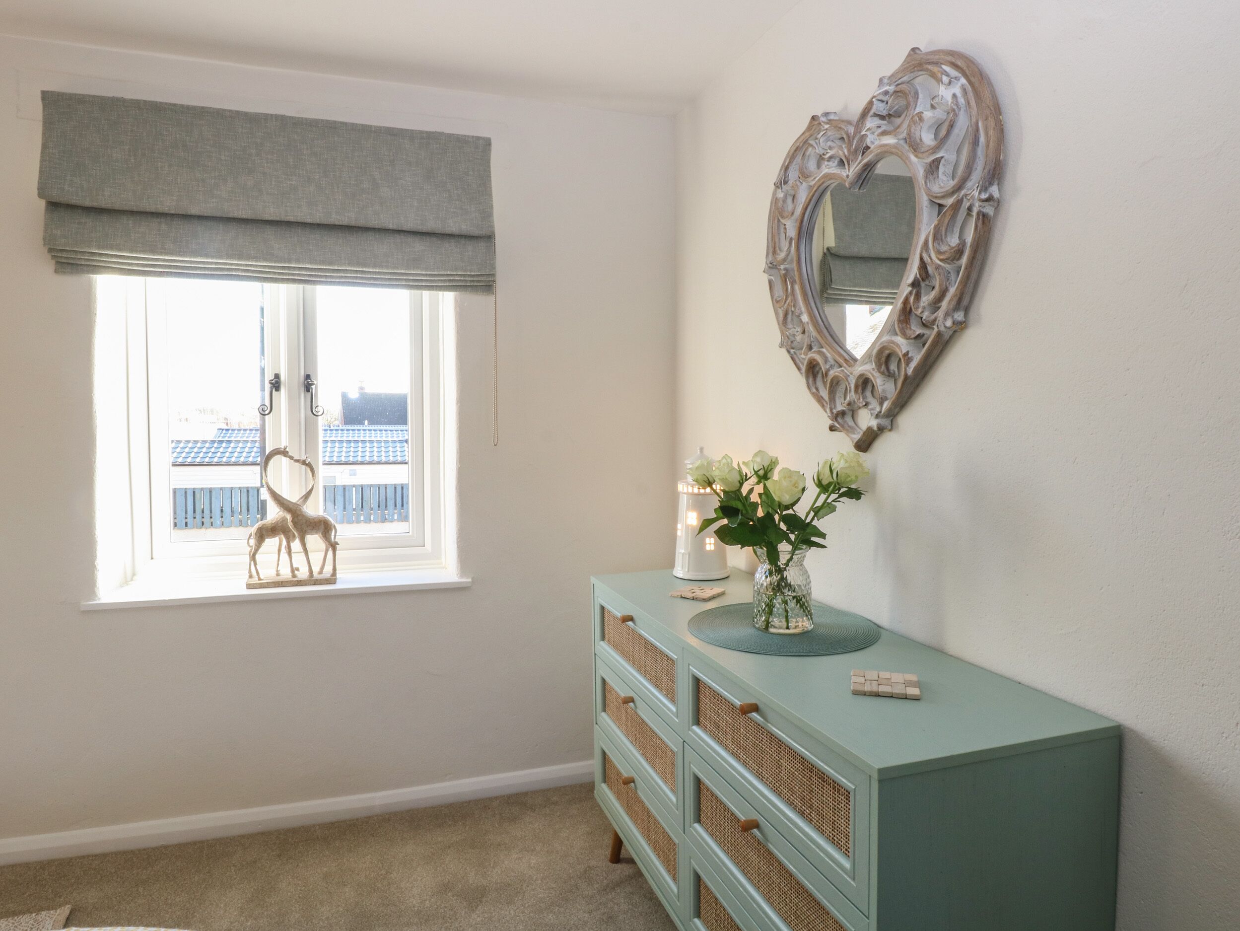 A room with a window, a dresser, a mirror, and a vase with flowers.