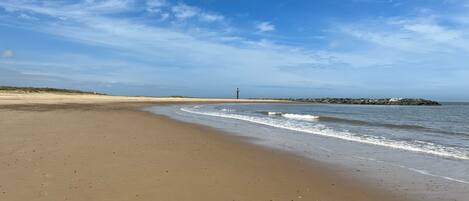 A wide sandy beach with gentle waves, a lighthouse in the distance, and a rocky outcrop on the right.