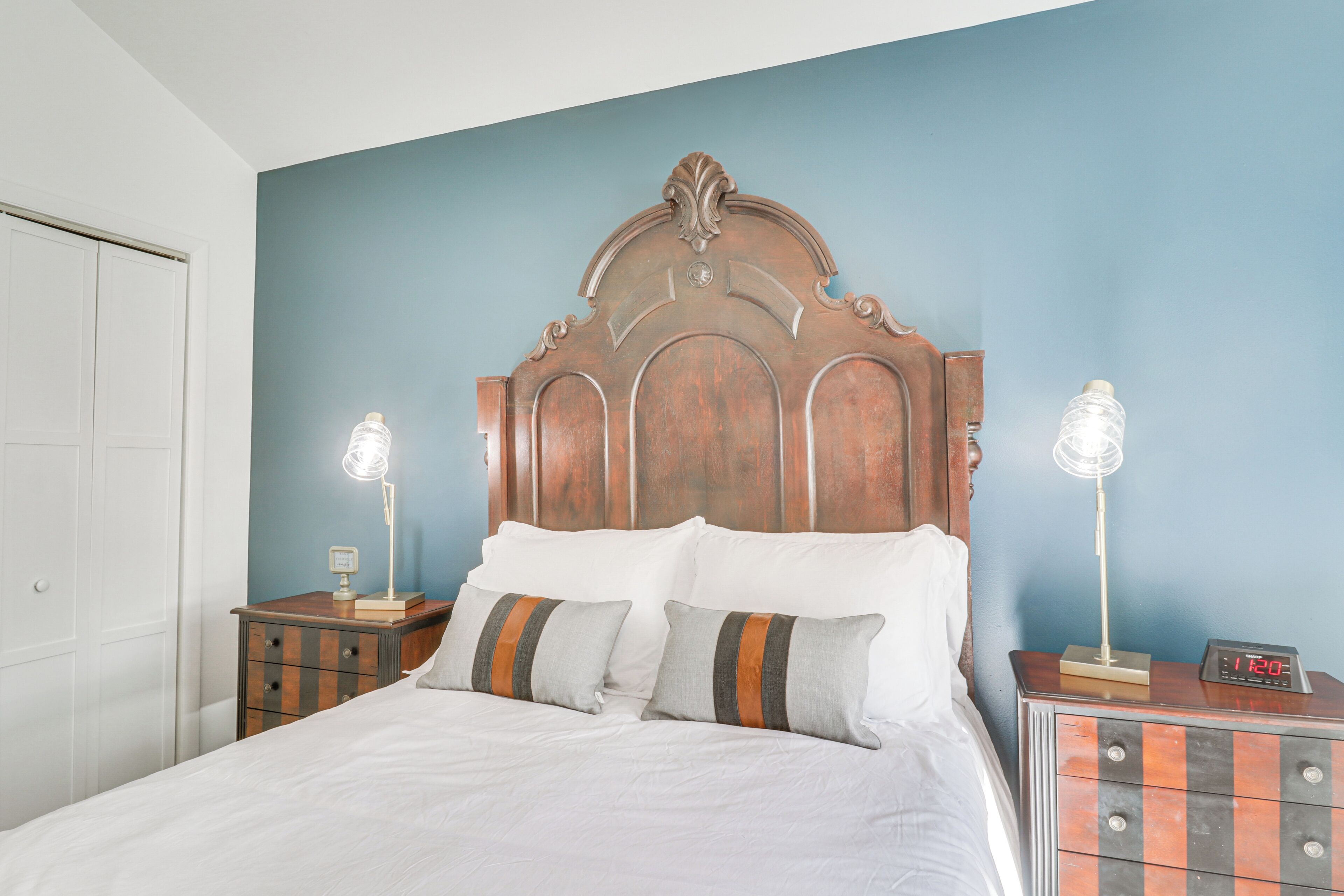 A bedroom with a wooden headboard, a bed with white bedding, two bedside tables with lamps, and a clock.
