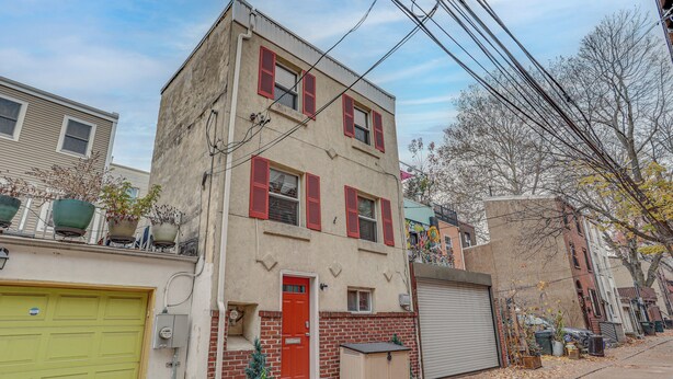 A two-story building with a red door, a green garage door, and a small front yard with a potted plant. - Centrally Located Philadelphia Townhome w/ Patio! (Philadelphia)