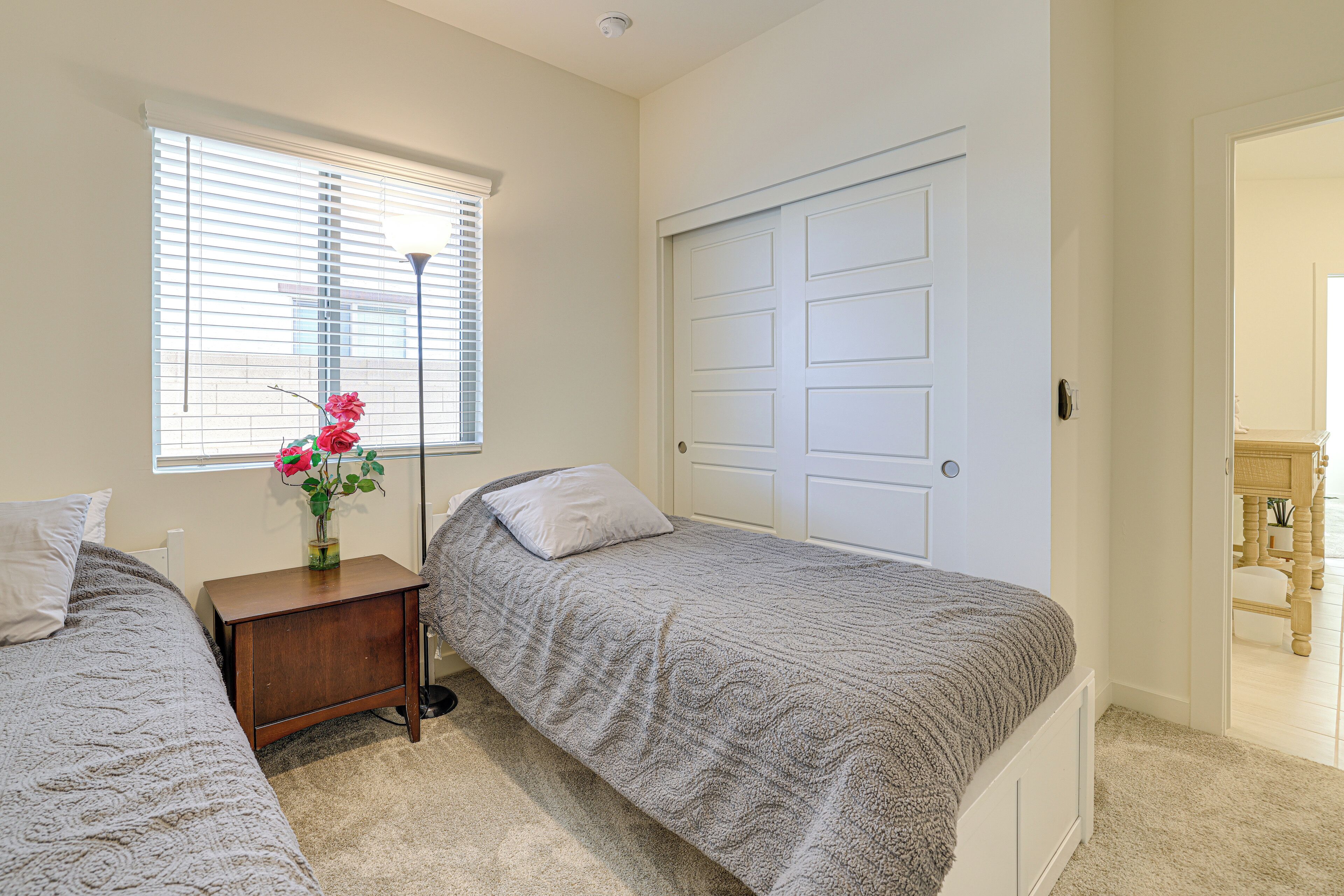 A bedroom with two single beds, a bedside table with a vase of flowers, a window with blinds, and a white door.