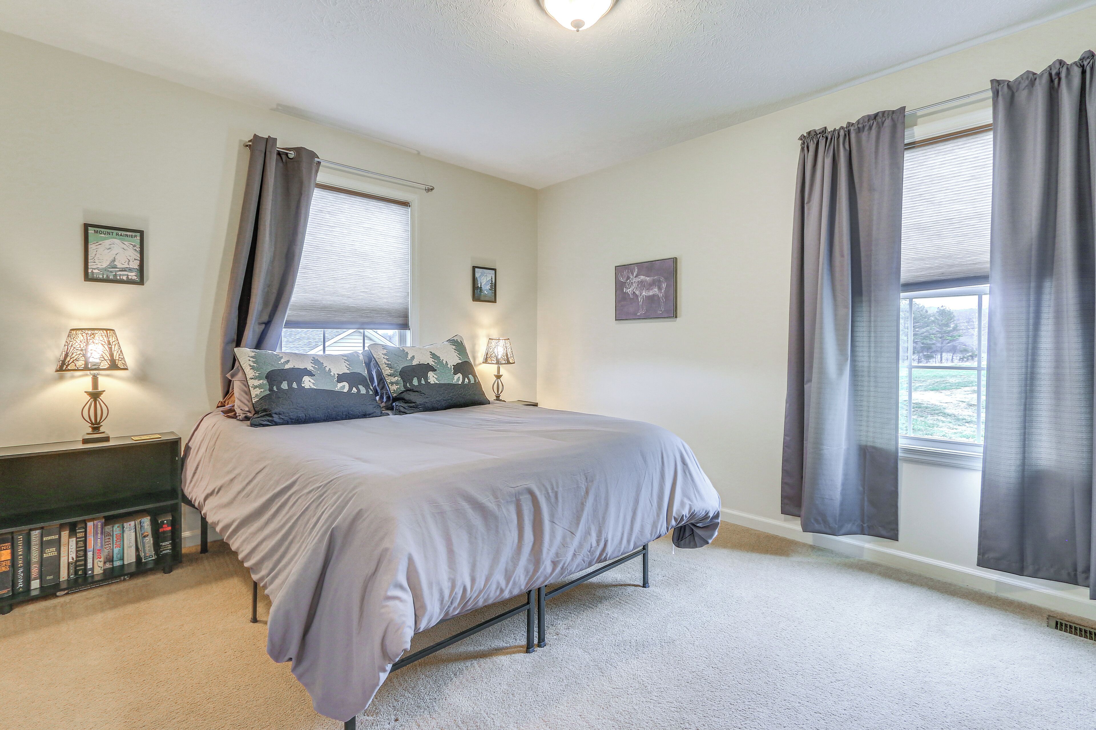 A bedroom with a bed, two bedside lamps, a bookshelf, and two windows with curtains.