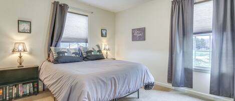 A bedroom with a bed, two bedside lamps, a bookshelf, and two windows with curtains.