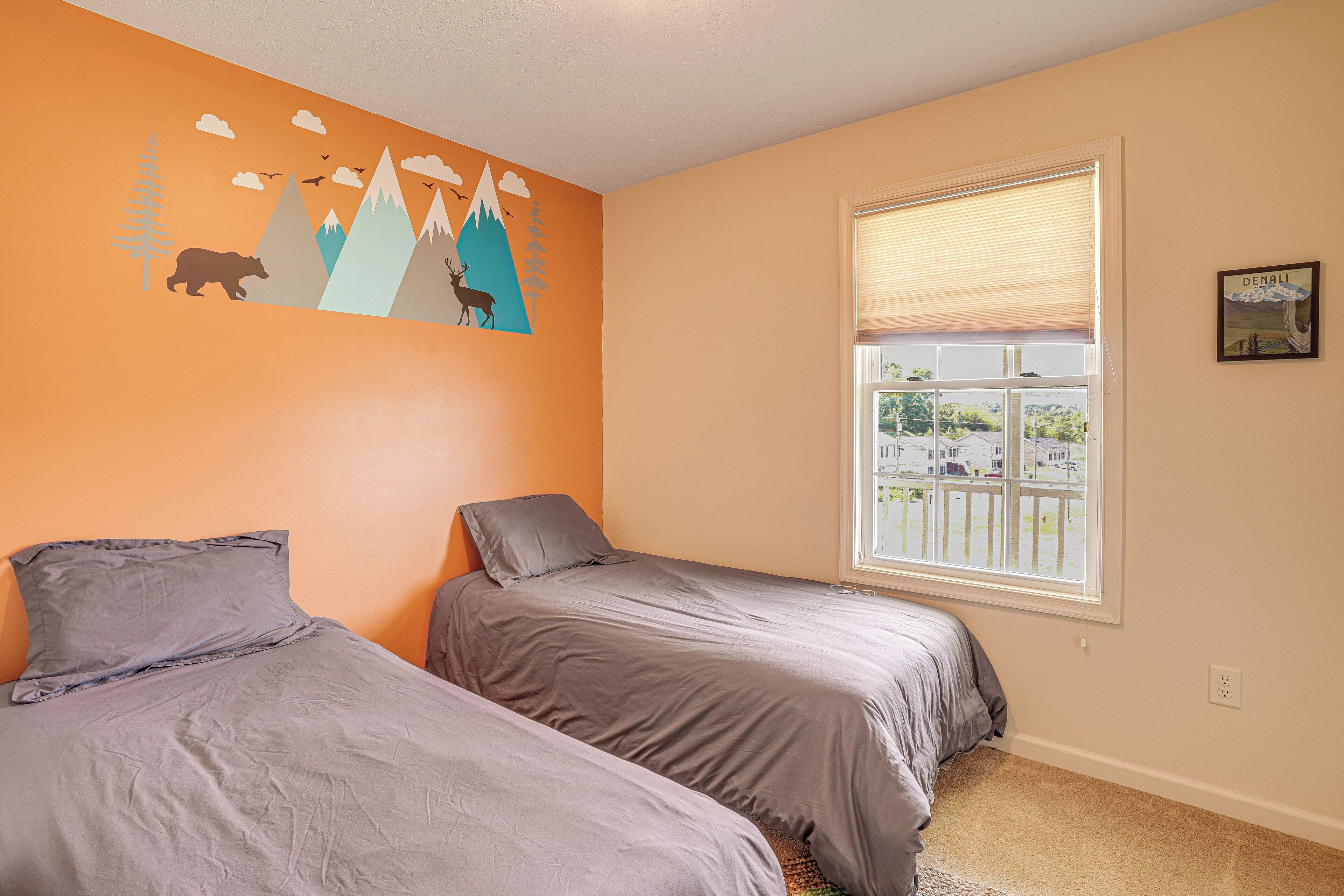 A bedroom with two beds, a mountain-themed wall mural, a window with blinds, and a small framed picture on the wall.