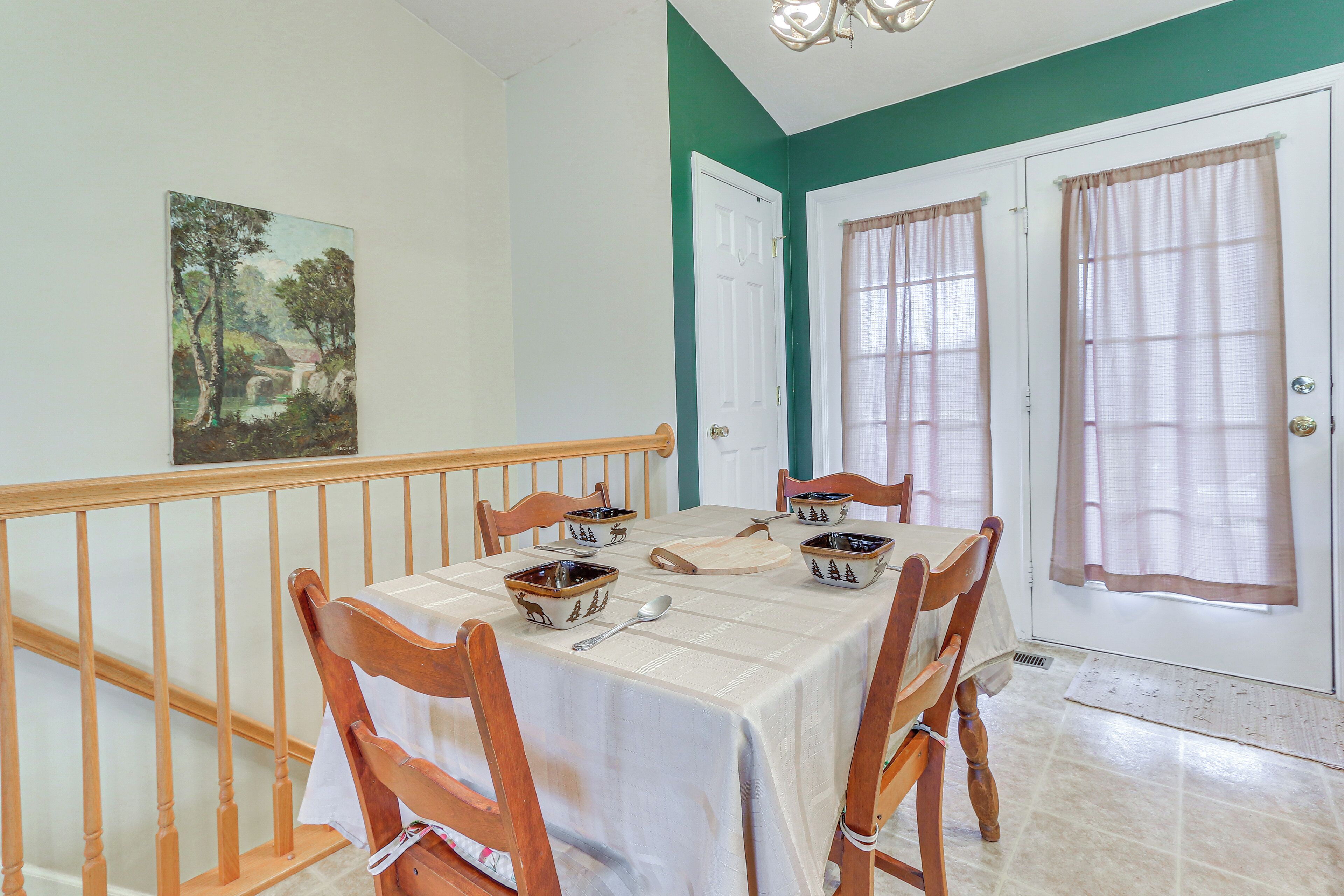 A dining area with a table set for four, wooden chairs, a painting on the wall, and a staircase with a railing.