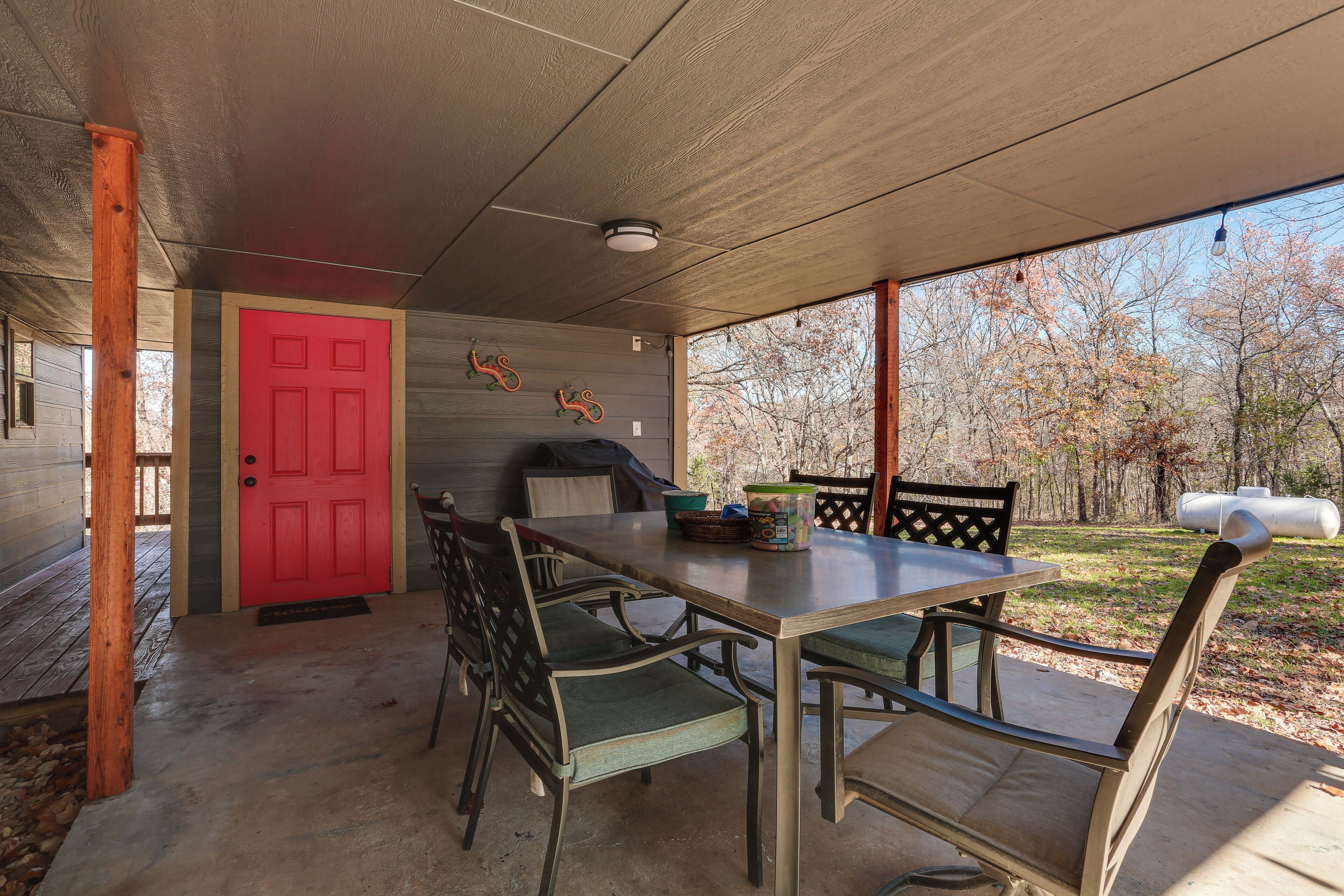 Un área de comedor al aire libre techada, con una puerta roja, sillas de madera y una mesa.