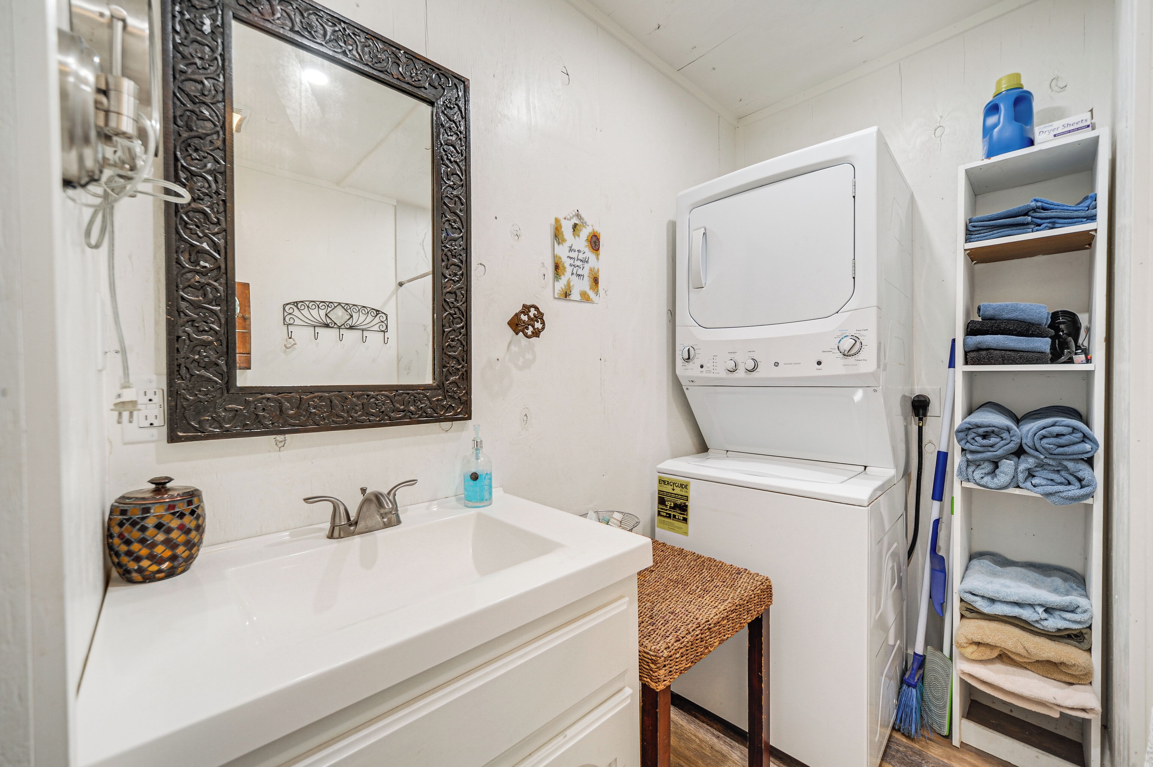 A compact bathroom with a white sink, a large mirror, a washing machine, and a shelf with folded towels.