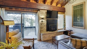 A cozy living room with a stone fireplace, wooden beams, and a view of the outdoors. - Mountain-view Condo in Brian Head w/ Private Sauna (Brian Head)