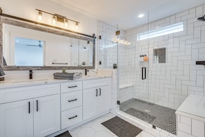 Uma casa de banho moderna com armários brancos, um espelho grande, chuveiro com portas de vidro e padrão de azulejos em padrão herringbone nas paredes.