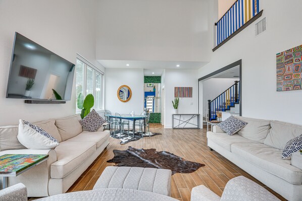 A modern living room with two sofas, a coffee table, and a TV mounted on the wall. There is a dining area with a table and chairs, and a staircase leading to another level.