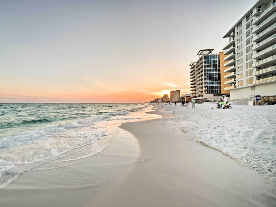 A beach with a sandy shore and ocean waves, adjacent to a multi-story hotel with balconies and a clear view of the sunset.