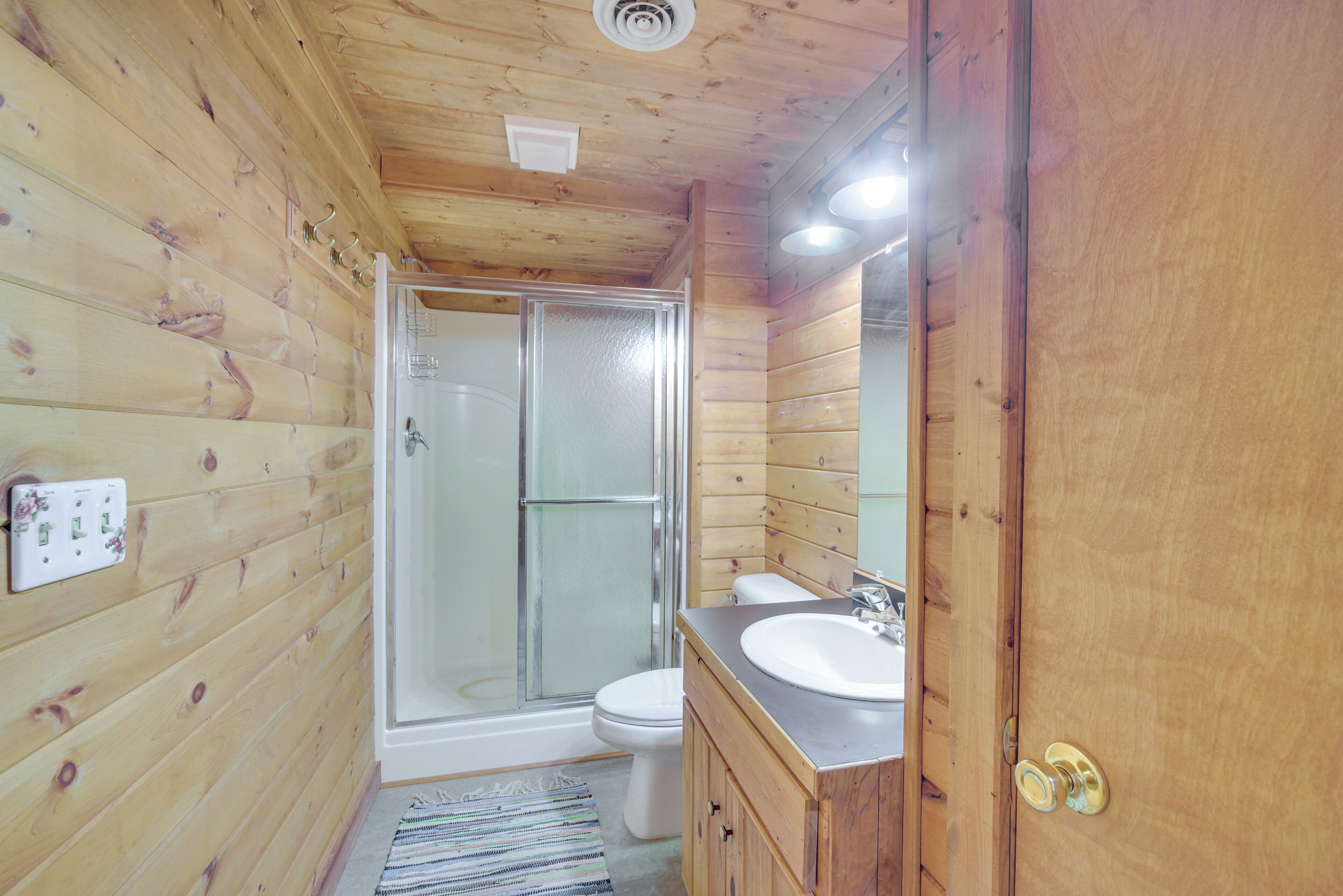 Un baño revestido de paneles de madera con una ducha envuelta en vidrio, un inodoro blanco y un tocador de madera con un lavabo.