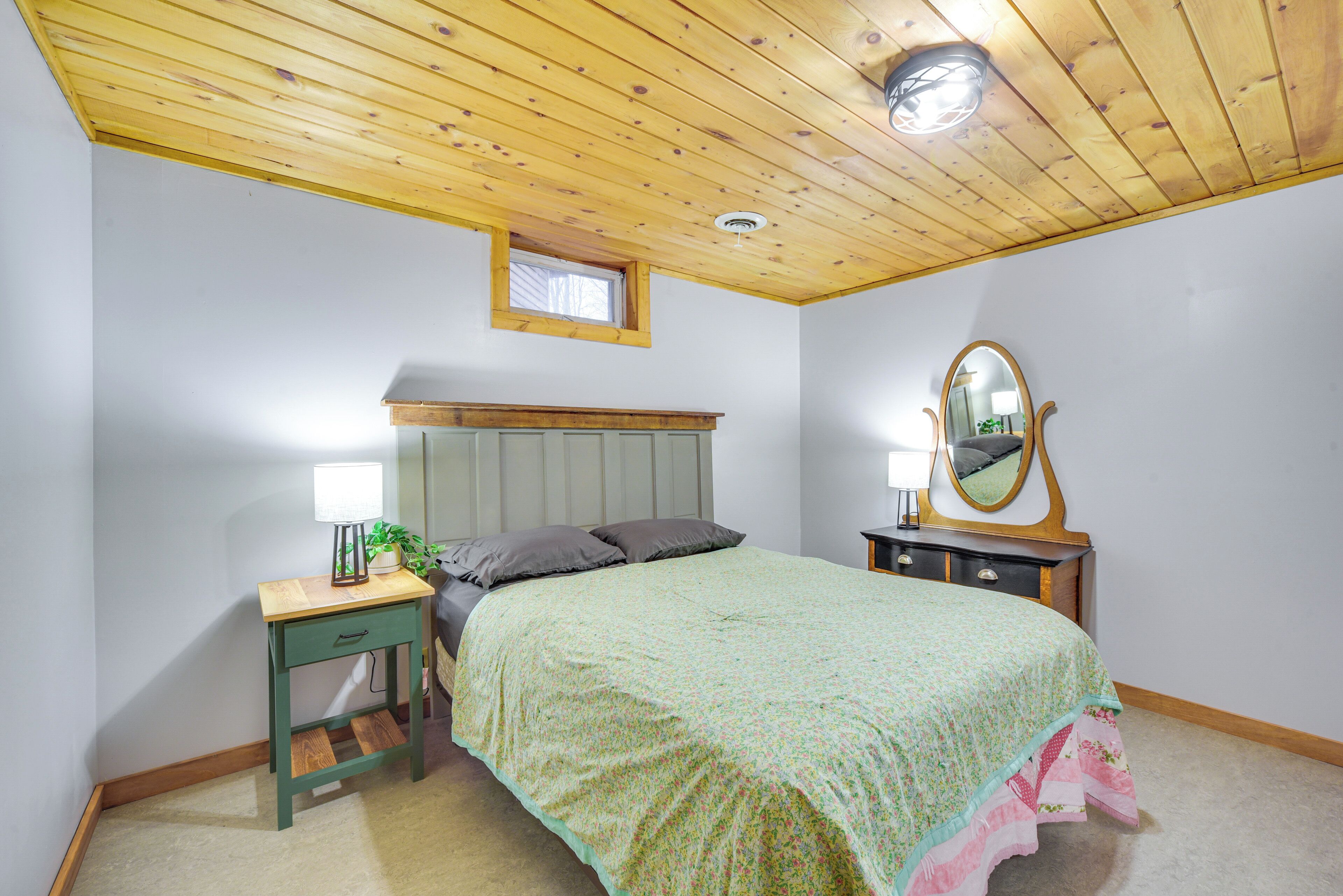 Une chambre avec un plafond en bois, un lit recouvert d’une couvre-lit à motifs floraux, une table de chevet avec une lampe, une commode avec un miroir et une petite fenêtre.