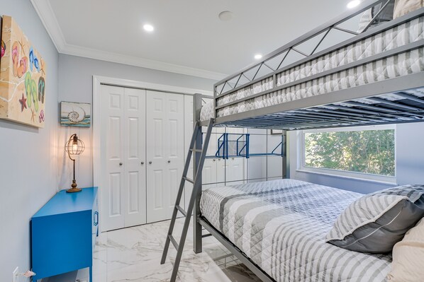 Una cama litera con escalera, un escritorio con lámpara, una puerta y una ventana con vista a árboles.