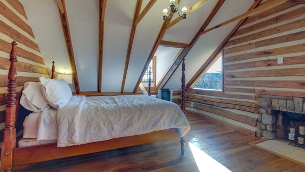 Une chambre avec un lit en bois, des poutres inclinées au plafond, une cheminée en pierre et une fenêtre.