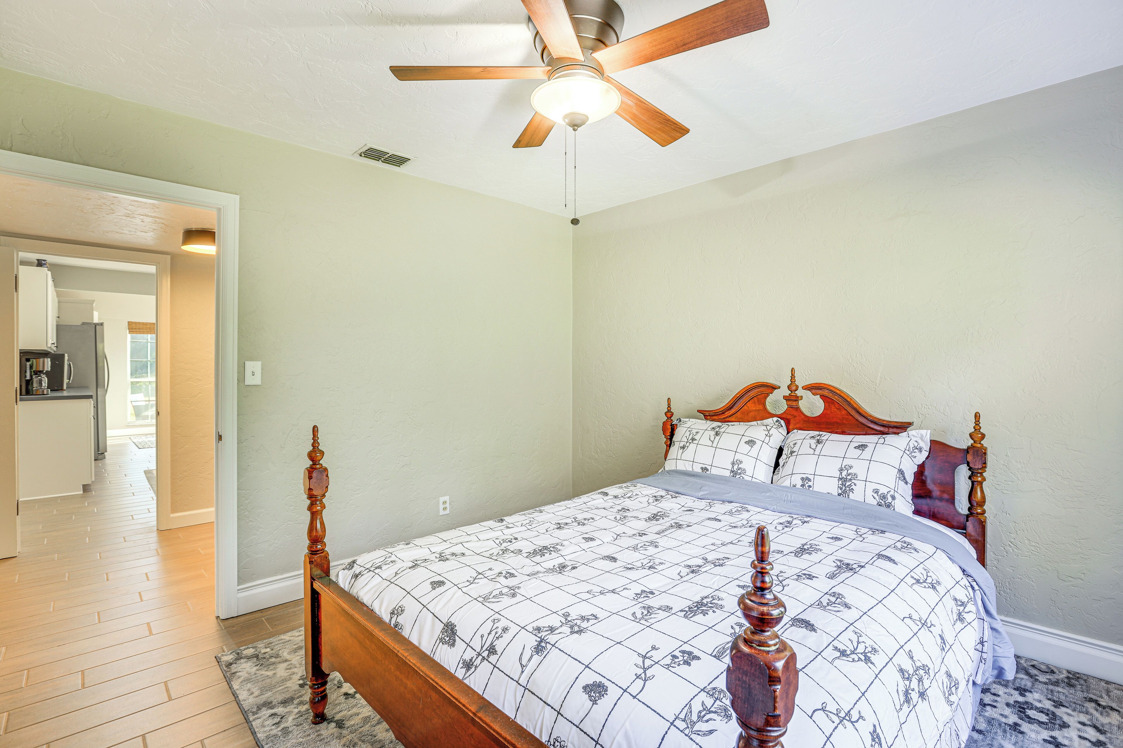 A bedroom with a wooden bed, a ceiling fan, and a view of a kitchen and hallway.