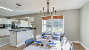 Un coin repas avec une longue table dressée pour six personnes, des meubles blancs et, au fond, une cuisine. Une grande fenêtre est agrémentée de stores.