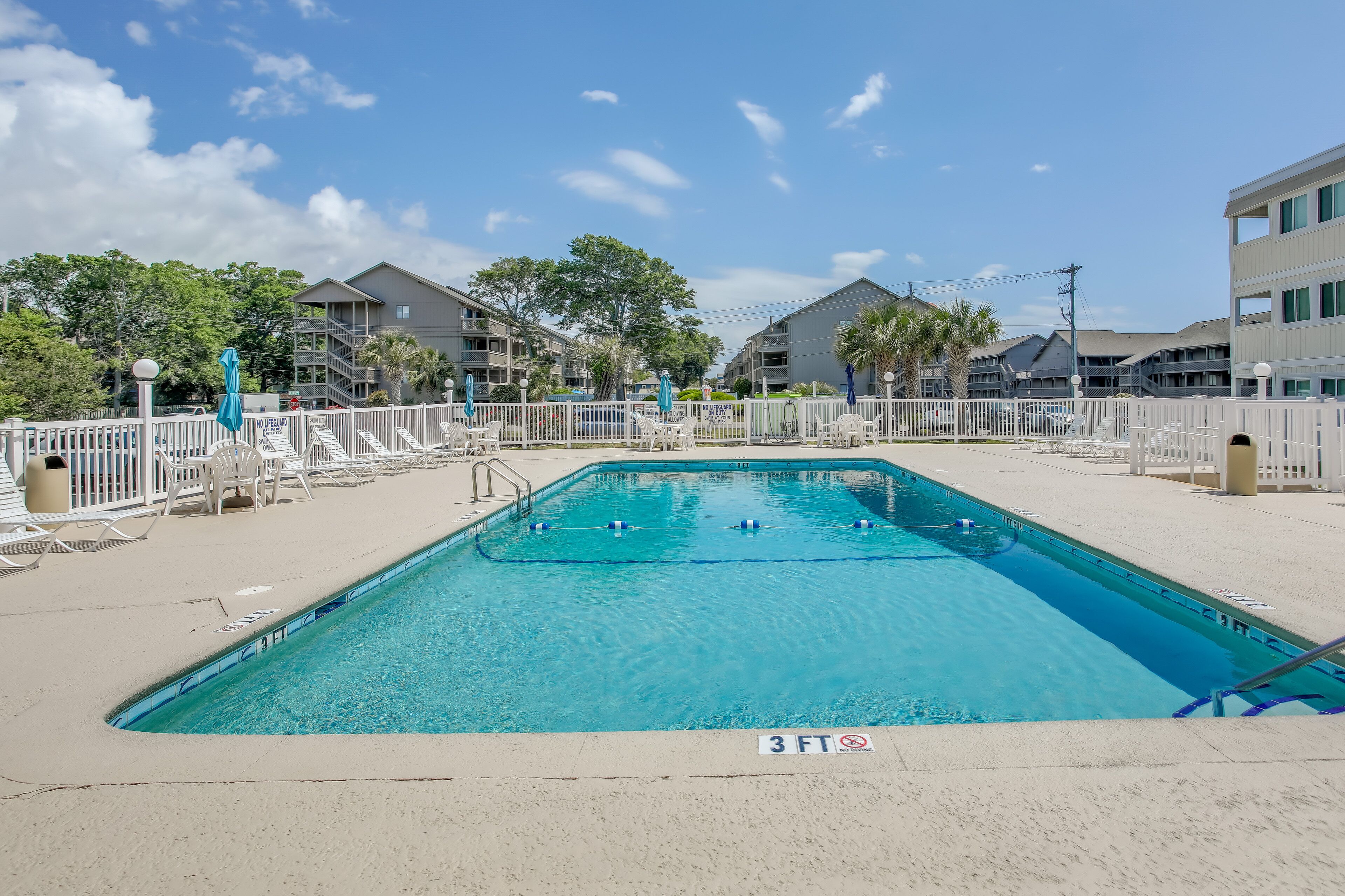 A rectangular swimming pool with a 3 FT depth marker, surrounded by a white fence, with buildings and trees in the background.