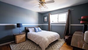 Une chambre à coucher comprenant un lit, une table de chevet, une lampe, une fenêtre avec des rideaux et un ventilateur de plafond.