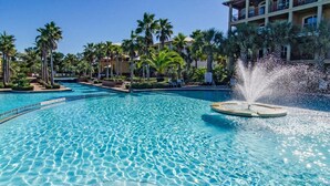 A clear blue pool with a water fountain, surrounded by palm trees and a building with balconies.