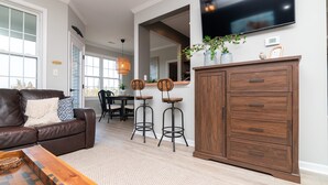 Un salon moderne avec un canapé en cuir marron, une table basse en bois et un téléviseur fixé au mur. On aperçoit deux tabourets de bar et une table à manger au fond de la pièce.