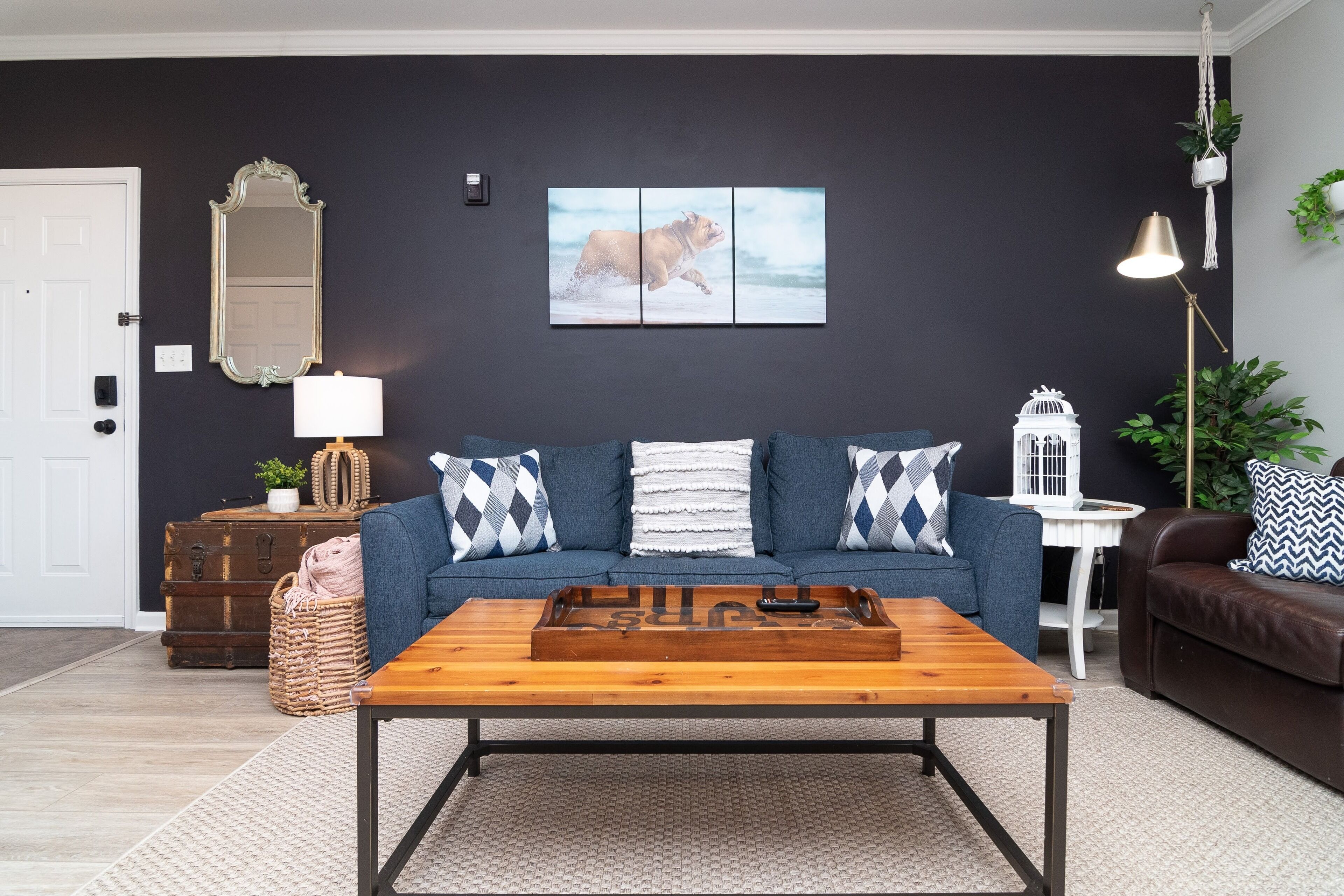 Una sala de estar con una pared oscura, una puerta blanca, un sofá azul con cojines estampados, una mesa de centro de madera y una mesita auxiliar blanca con una lámpara.