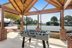 Una mesa de fútbol de mesa en un área exterior cubierta, con pilares de ladrillo y una cancha de tenis al fondo.