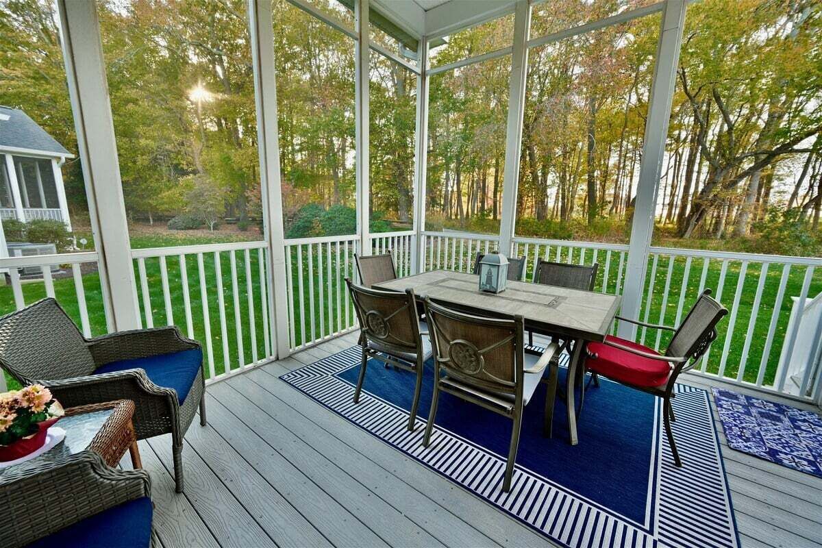 A covered outdoor deck with a table and chairs, surrounded by a white fence and trees.