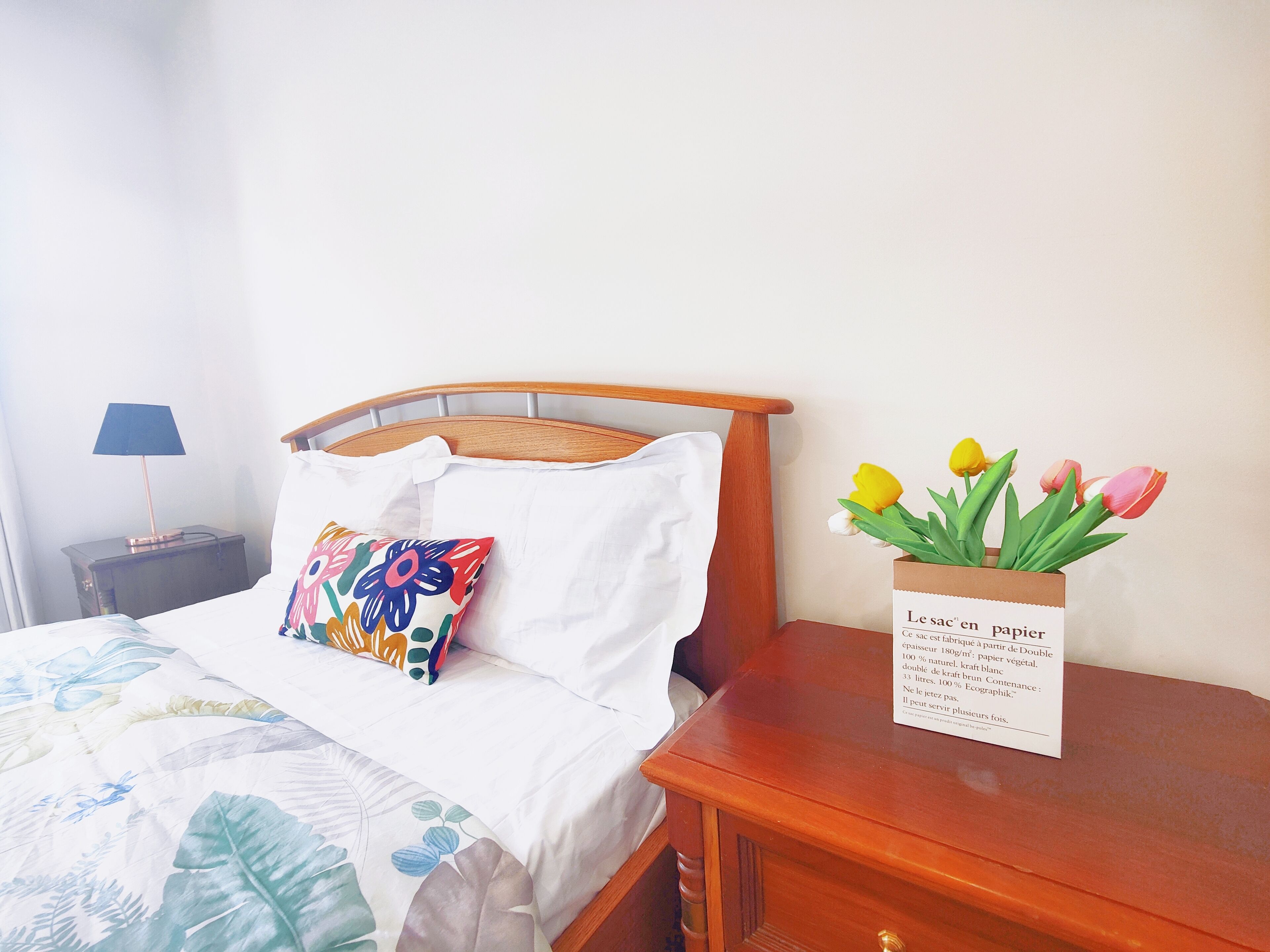 Un lit bien fait, agrémenté d’un coussin à motifs floraux et d’un coussin blanc, une table de chevet avec une lampe et un vase de tulipes posé sur une table de nuit en bois.