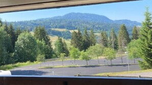 Blick aus einem Fenster mit einer hölzernen Bank im Vordergrund, auf eine Straße und eine bewaldete Landschaft mit Bergen in der Ferne.