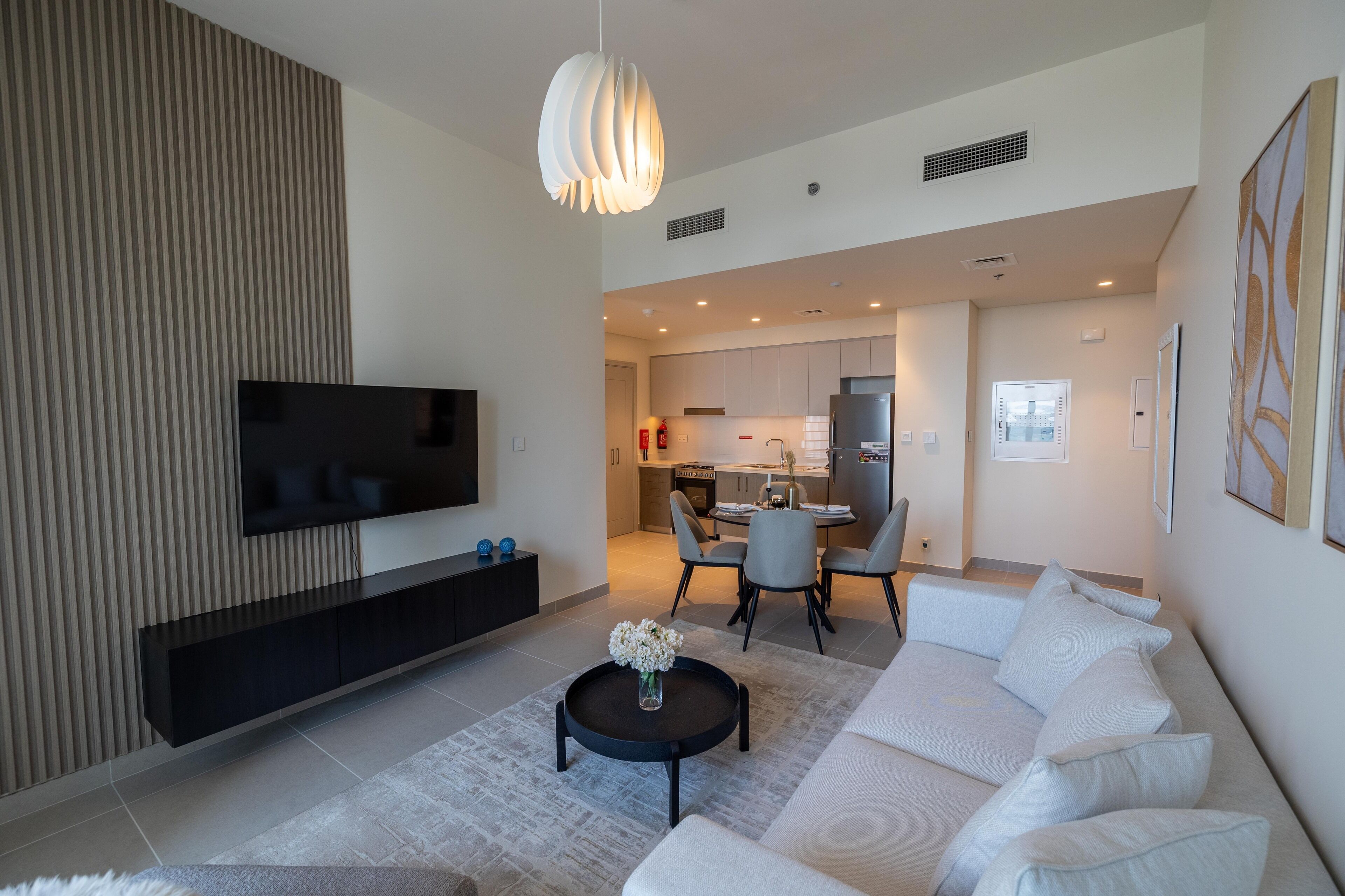 Een moderne woonkamer met een flatscreen televisie, een zwarte salontafel met bloemen, een witte bank en een eethoek met stoelen en een tafel.