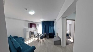 A modern living room with a blue sofa, a dining area with a table and chairs, and a kitchen area with a refrigerator and cabinets. - BBC Apartments (Navodari)