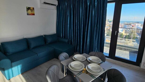 A modern living room with a sectional sofa, a dining table set for four, and a large window with a city view. - BBC Apartments (Navodari)