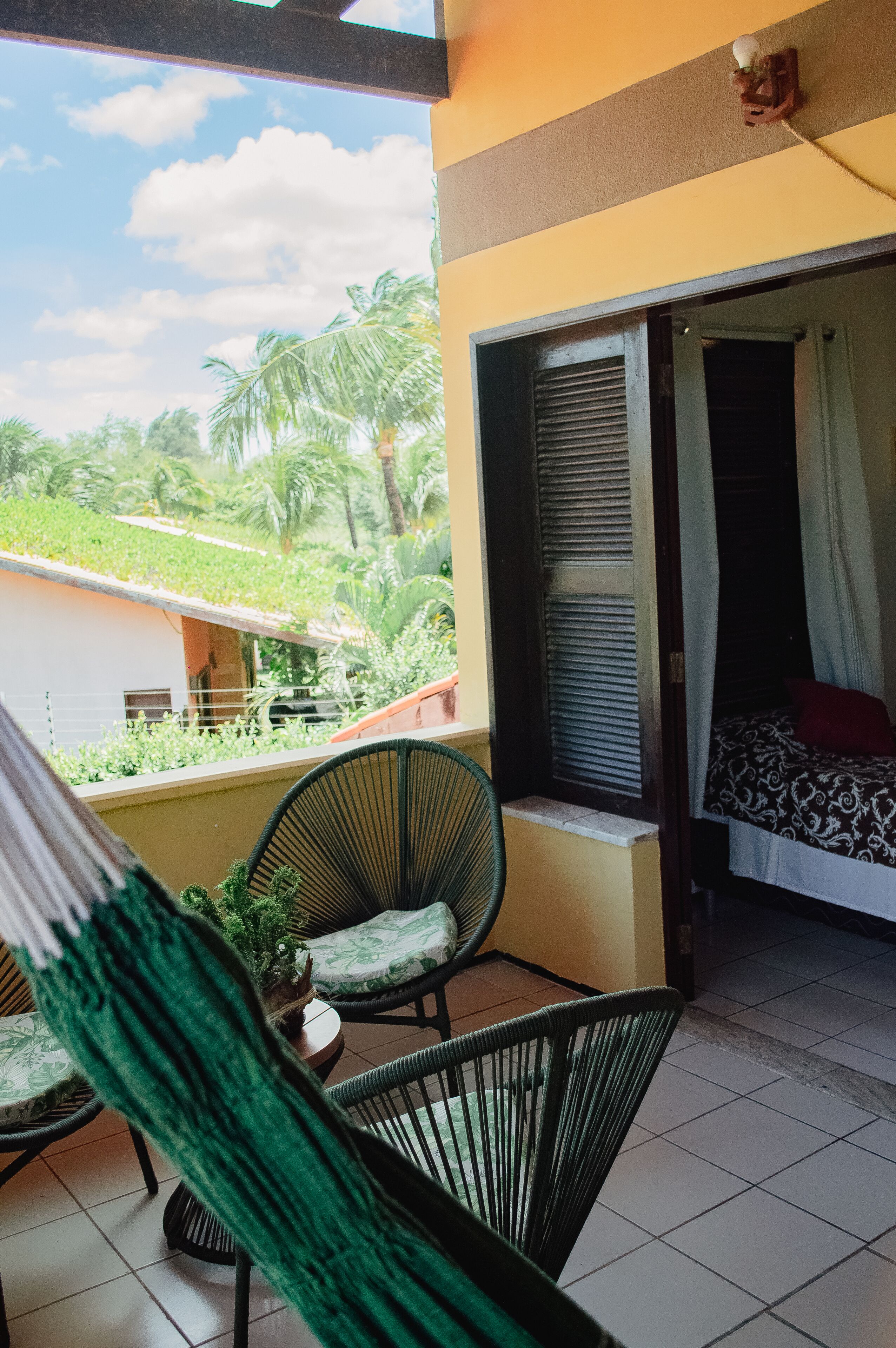 Un balcón con vistas a zonas verdes y un edificio, una cama con colcha estampada, y una silla con cojín.