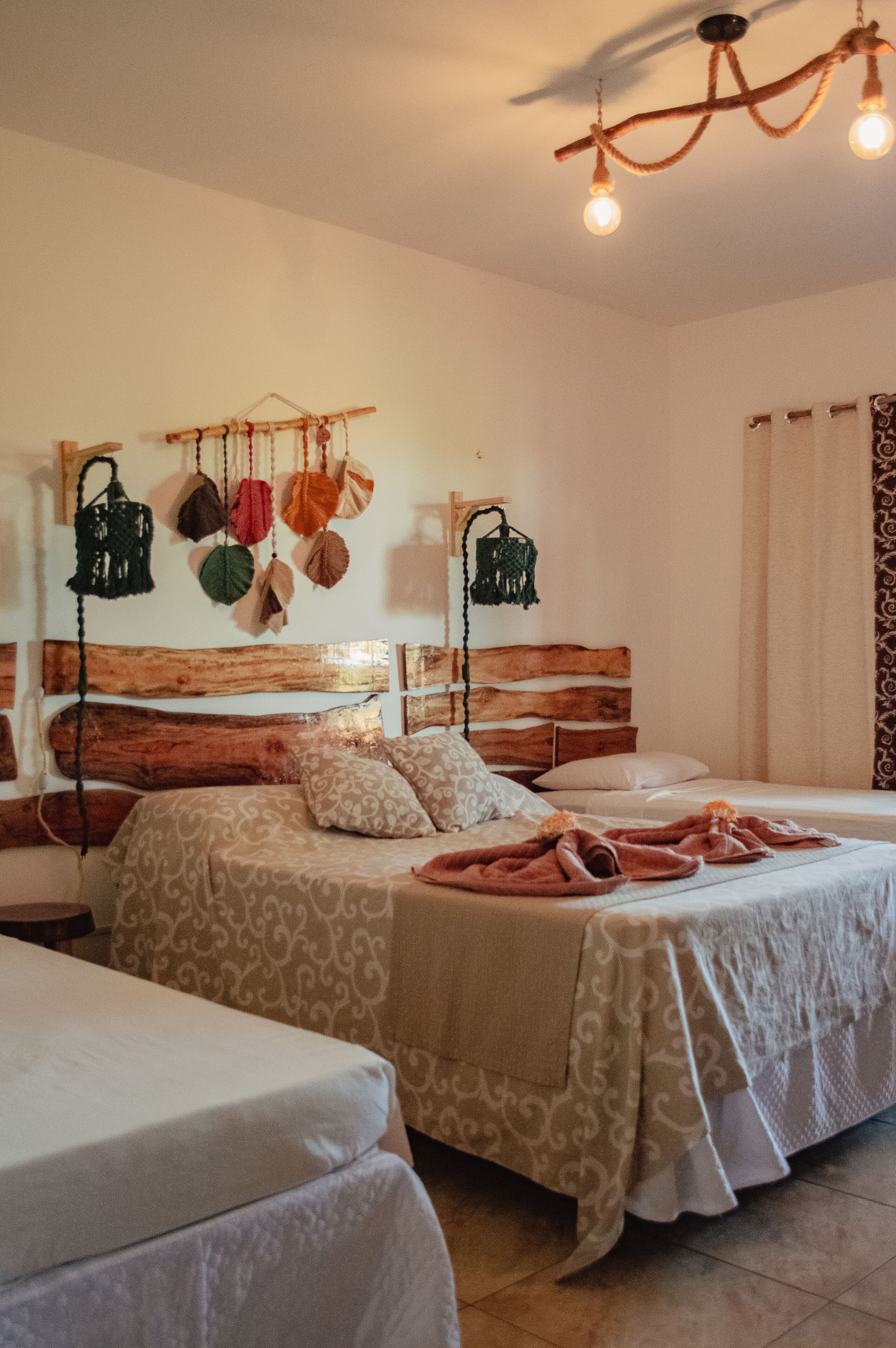 A cozy bedroom with a wooden headboard, two beds with patterned bedding, a hanging macrame wall decoration, and a ceiling light fixture.