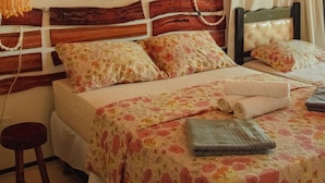Ein Schlafzimmer mit Holz-Kopfteil, einem Bett mit floralem Bettbezug, einer hängenden Makramee-Wanddekoration und einer Wand-Klimaanlage.