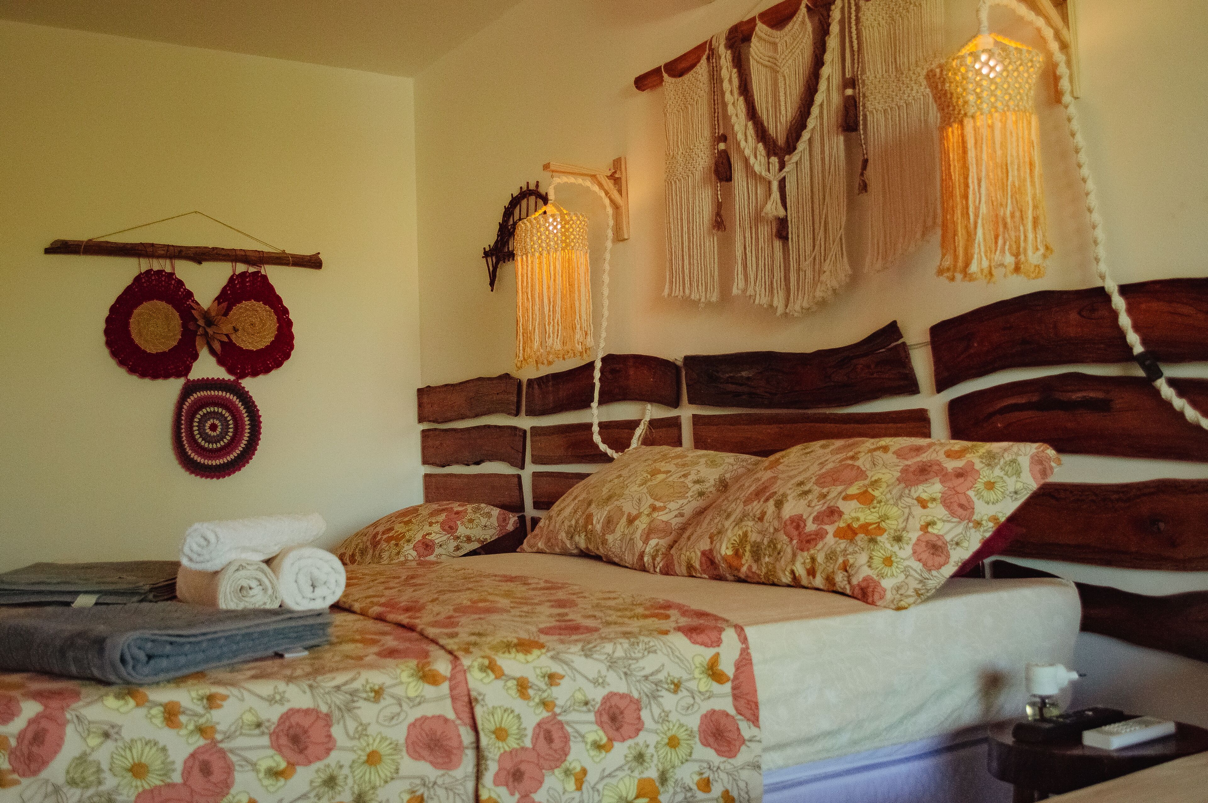A bed with floral bedding, a wooden headboard, and a wall-mounted macrame light fixture.