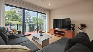 Una sala moderna con un televisor de pantalla plana grande sobre un mueble de madera, sofás grises, una mesa de centro y un balcón con vistas a zonas verdes.