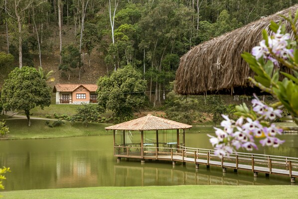 Un quiosco con techo de paja sobre una plataforma junto al lago, una casa con tejado de tejas al fondo y una ladera boscosa.