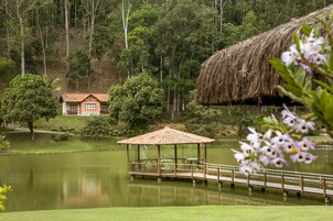 Un quiosco con techo de paja sobre una plataforma junto al lago, una casa con tejado de tejas al fondo y una ladera boscosa.