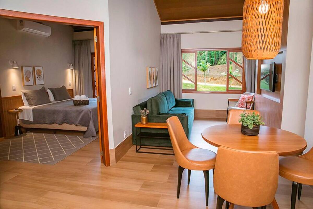 A modern room with a bed, a green sofa, a dining table with chairs, and a large window with a view of greenery.