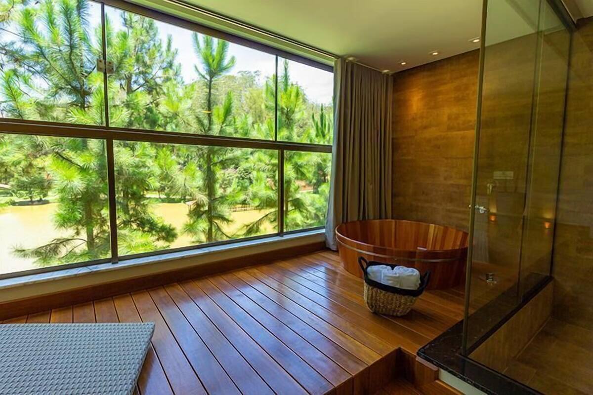 A modern bathroom with a large window, wooden flooring, a wooden bathtub, and a glass shower enclosure.