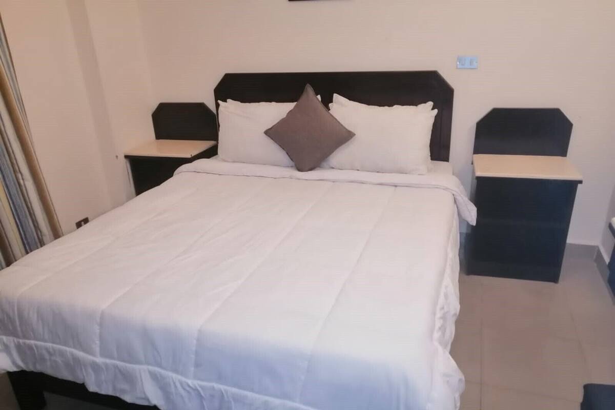 Een net opgemaakt bed met witte beddengoed en een bruin kussen, met aan weerszijden twee nachtkastjes met zwarte bovenkant en witte poten.