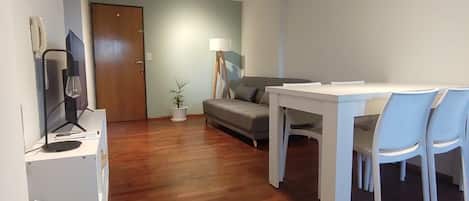 A modern living room with a wooden floor, a grey sofa, a white dining table with chairs, and a small side table with a lamp.