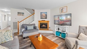 A living room with a fireplace, a staircase, and a TV displaying 'SEASIDE VACATIONS & SALES'.