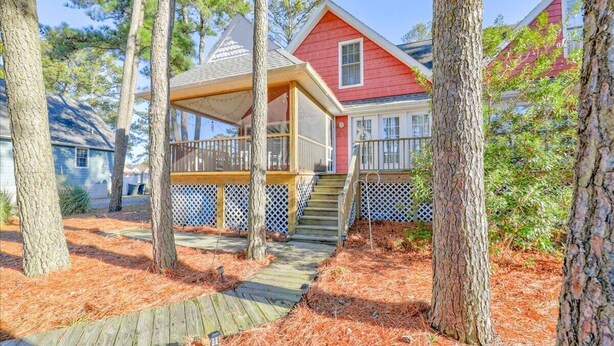 A wooden deck leading to a covered porch with a staircase, surrounded by pine trees and a red house.